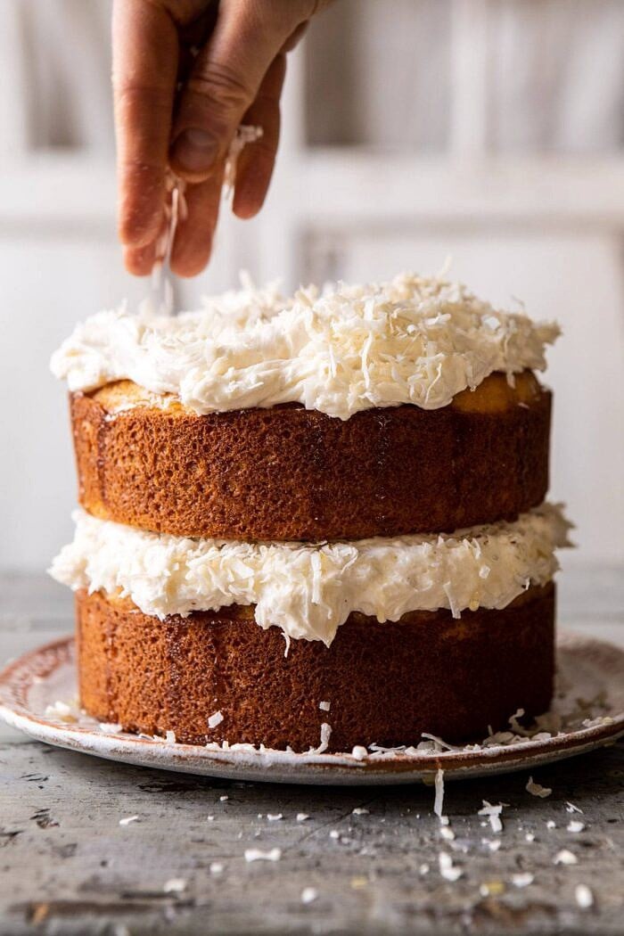Lemon Coconut Naked Cake with Whipped Vanilla Buttercream | halfbakedharvest.com #coconutcake #springrecipes #easter #cake #layercake Lemon Coconut Naked Cake with Whipped Vanilla Buttercream being sprinkle with coconut and hand in photo