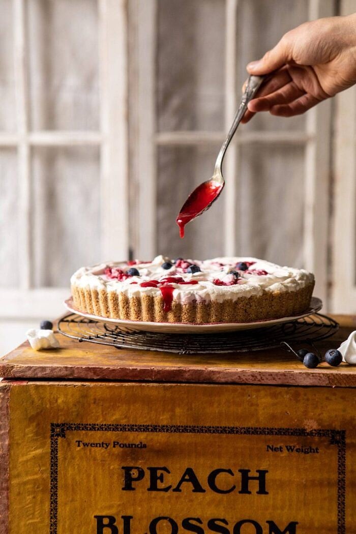 No-Bake Eton Mess Berry Cheesecake | halfbakedharvest.com #cheesecake #nonbake #easyrecipes #dessert #4thofjuly head on photo of No-Bake Eton Mess Berry Cheesecake with raspberry sauce being drizzled over cake